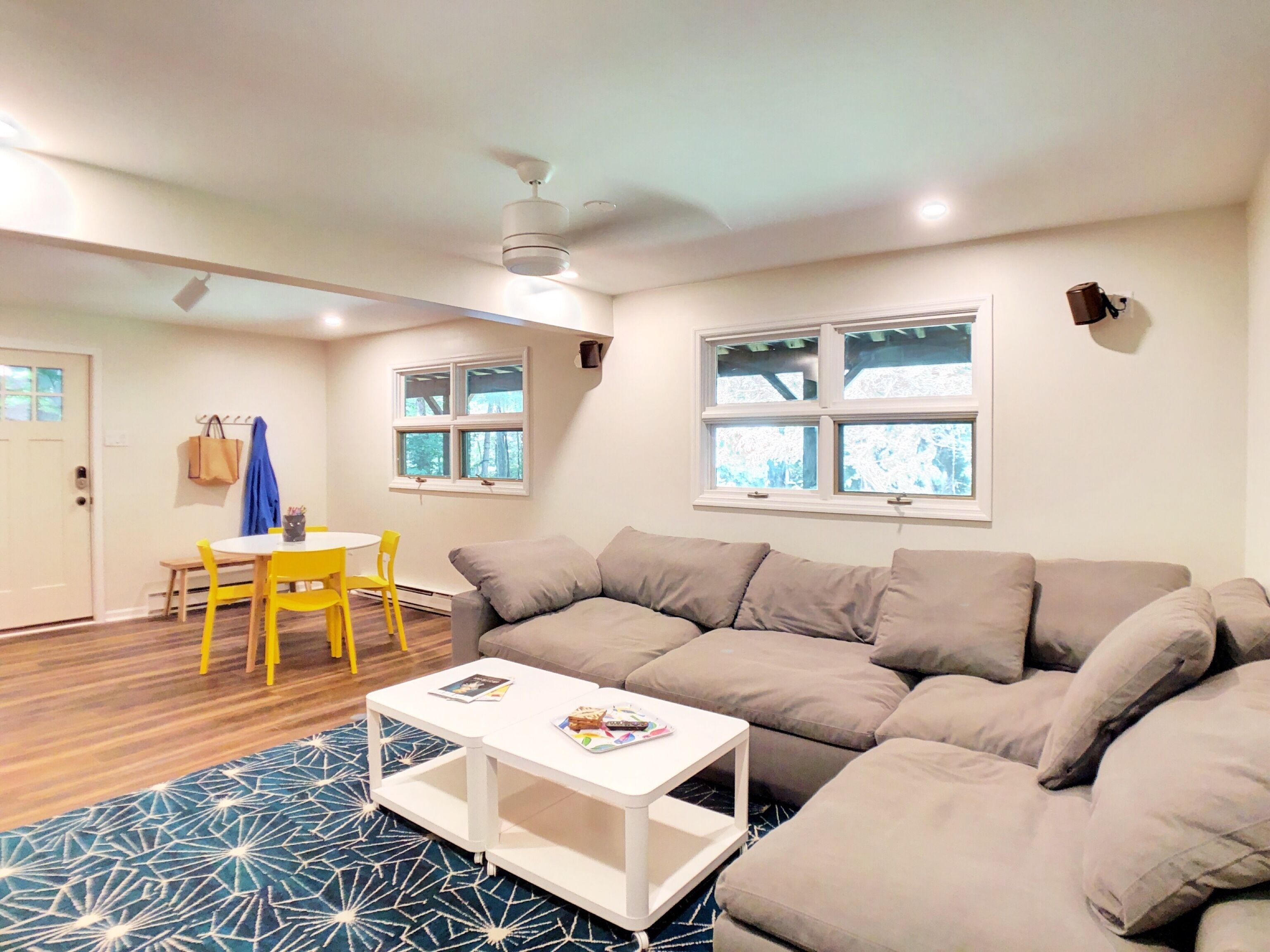 A living room with a grey sectional sofa, a white coffee table, a blue patterned rug, a dining table with yellow chairs, and two windows with white frames.
