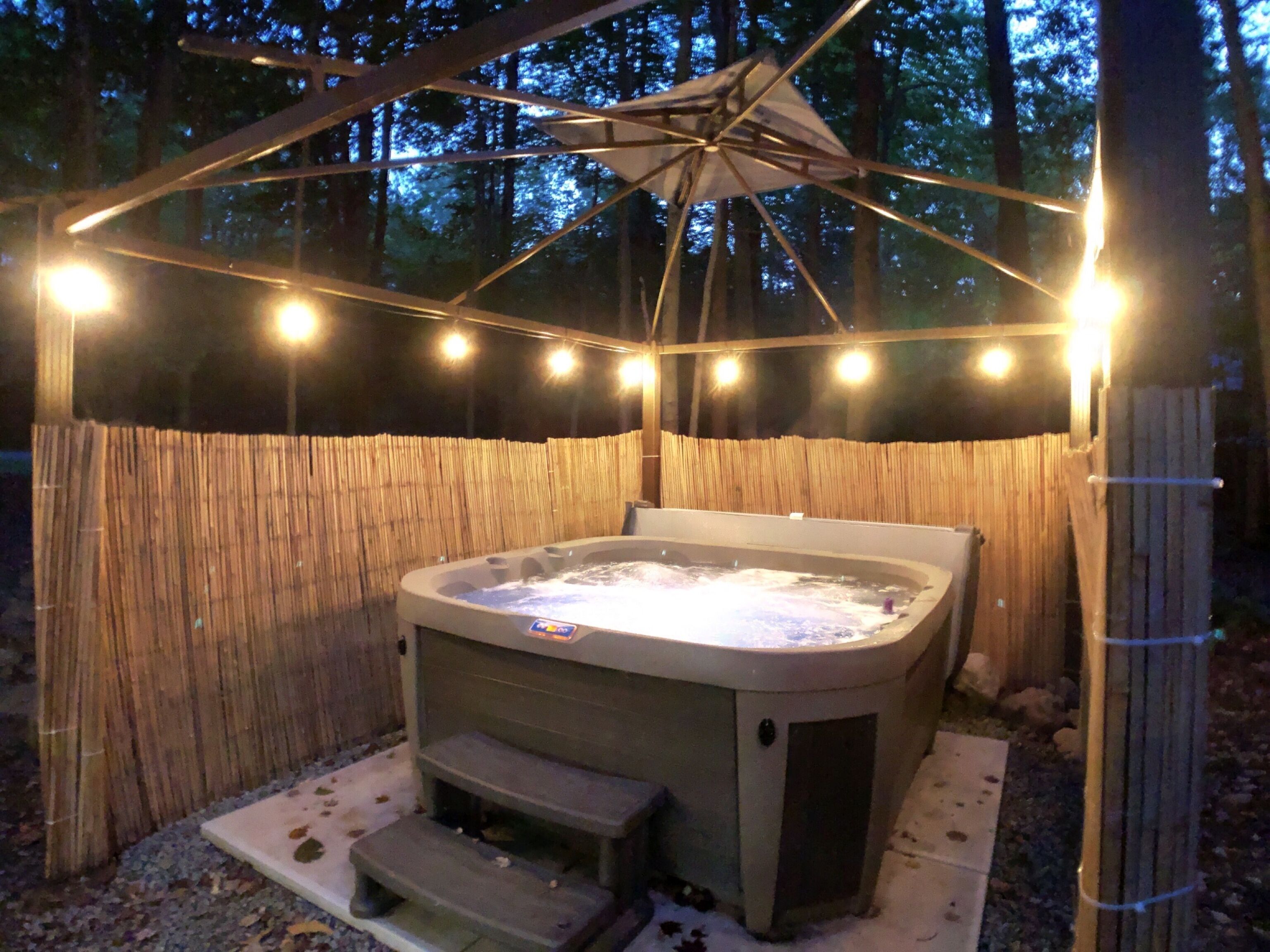 A hot tub with steps, surrounded by a bamboo fence, under a wooden pergola with string lights.