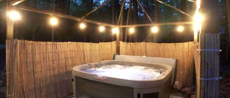 A hot tub with steps, surrounded by a bamboo fence, under a wooden pergola with string lights.