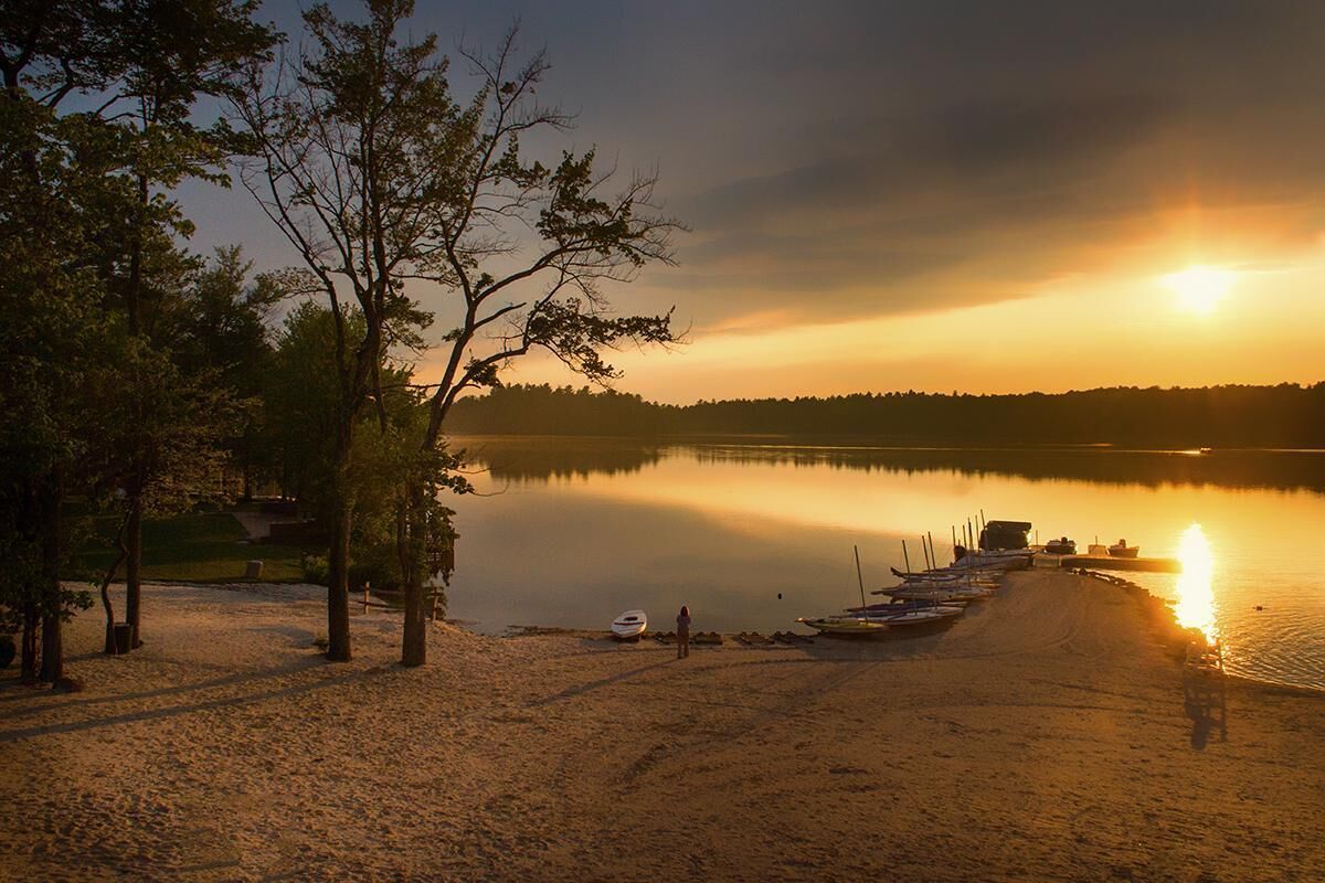 A lakeside scene with a sandy beach, trees, and boats docked along the shore.