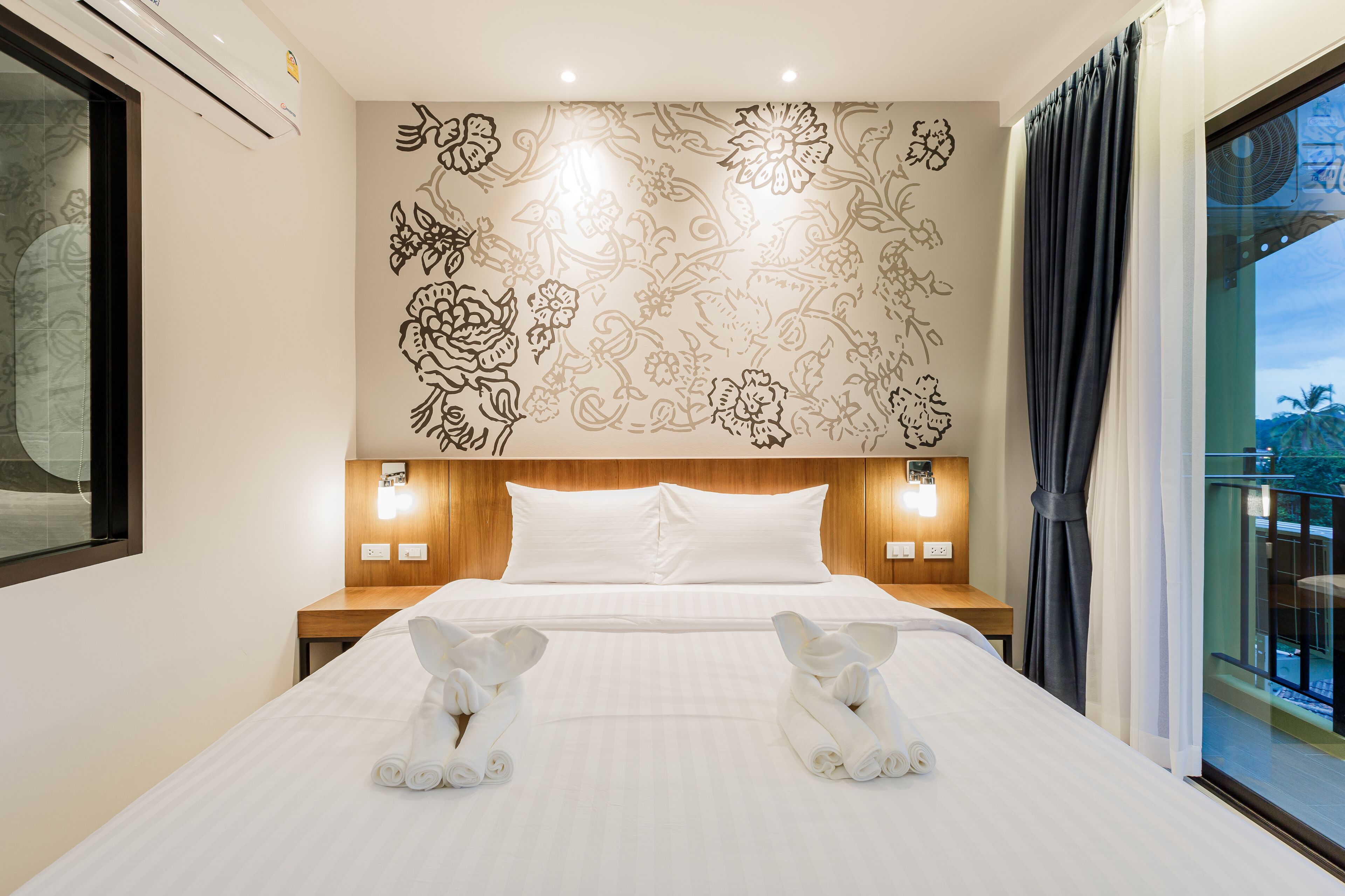 Quarto de hotel com uma cama grande, decoração floral nas paredes, luminárias de cabeceira e vista para o exterior.