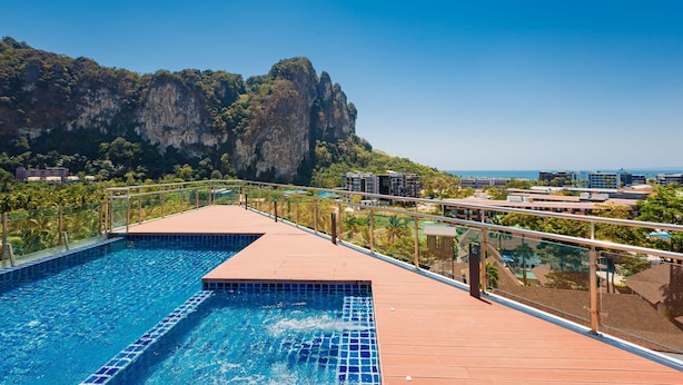 Une piscine dotée d’une terrasse en bois et d’une balustrade en verre, offrant une vue sur un paysage montagneux et sur la ville.