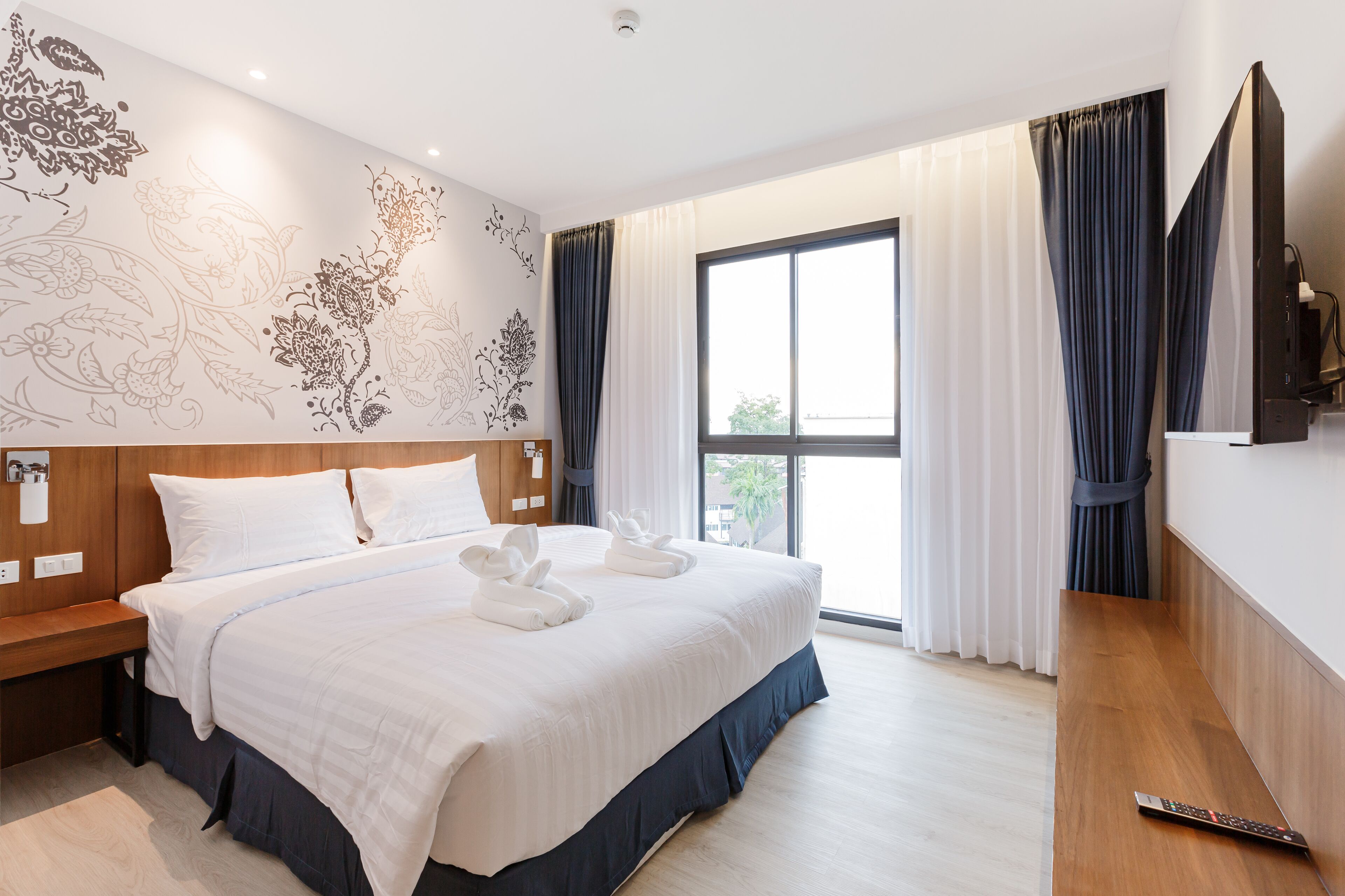A hotel room with a large bed, a wooden bench, a flat-screen TV, and a large window with curtains.