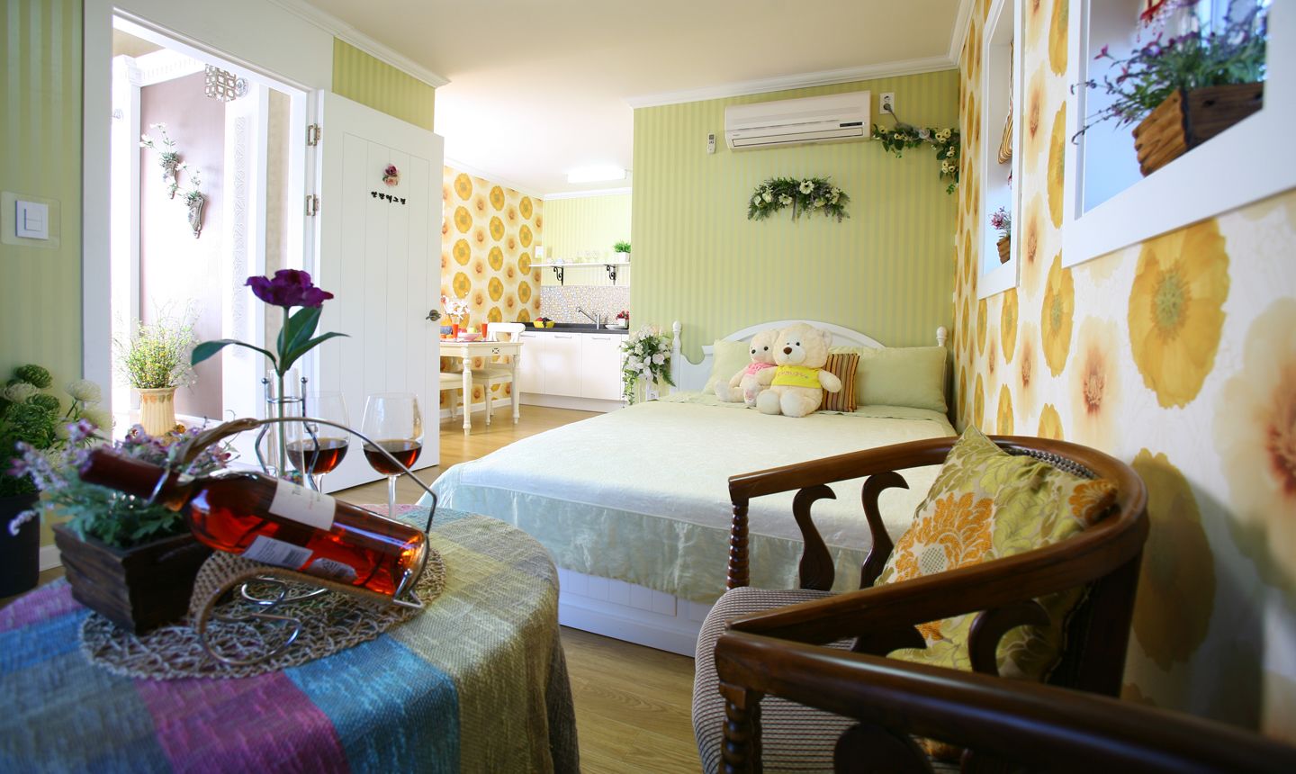A cozy bedroom with a bed, a wooden chair, a dining table with a vase of flowers, and a kitchen in the background.