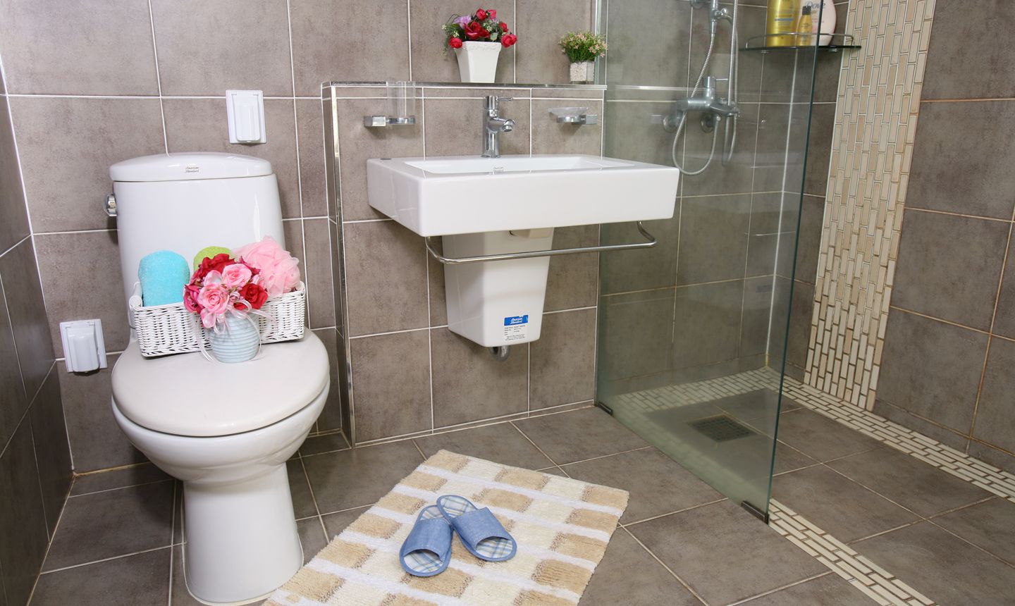 A bathroom with a toilet, sink, and shower. There are slippers on the floor, a towel rack, and a small plant on the sink.