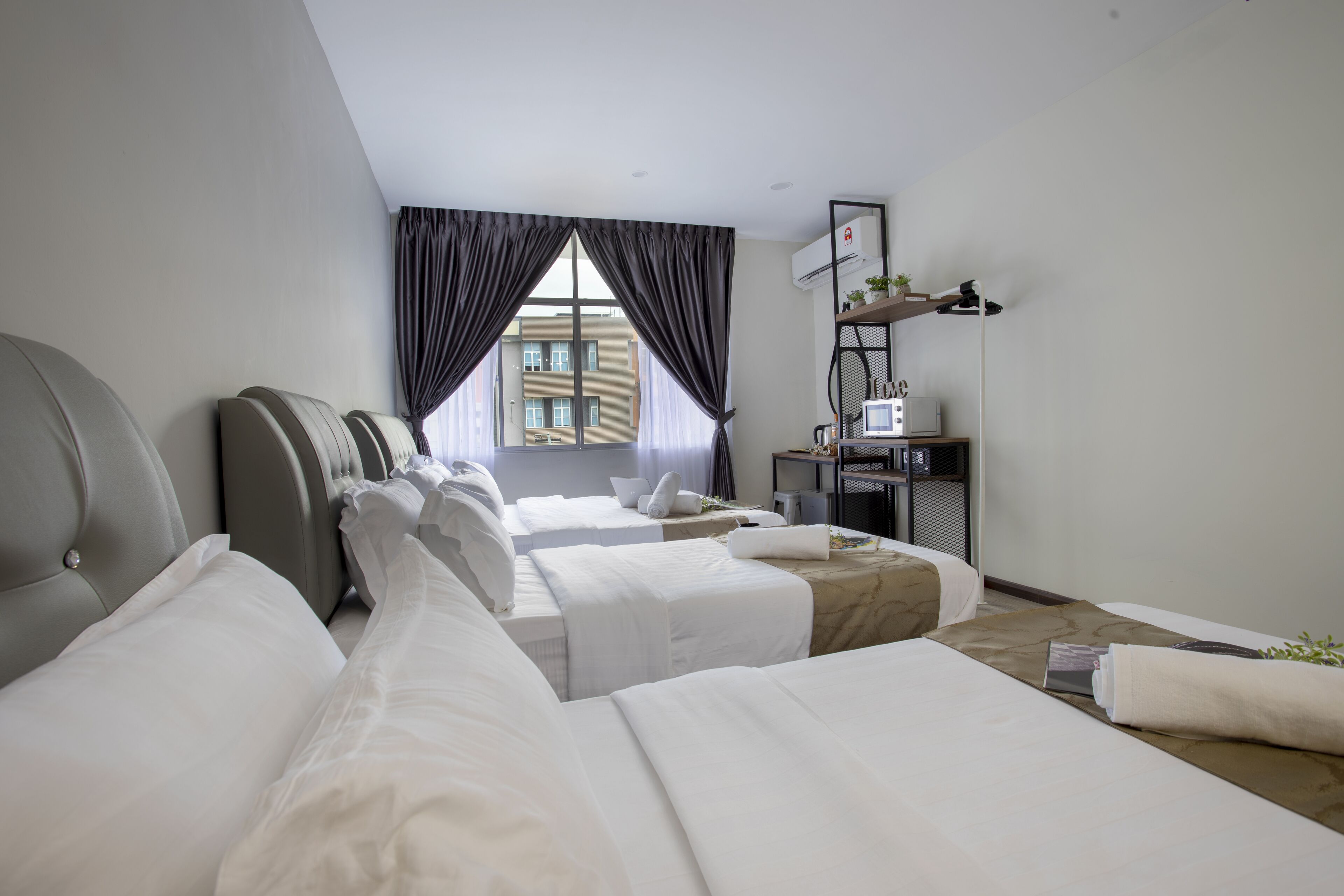 A hotel room with two beds, a desk, a chair, a window with curtains, and a small shelf with items.