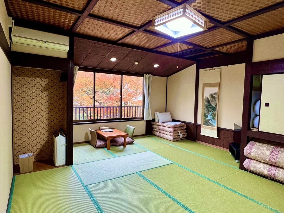 A traditional Japanese room with tatami flooring, a sliding door, a window with autumn foliage, a tatami mat table, and a low wooden bench.
