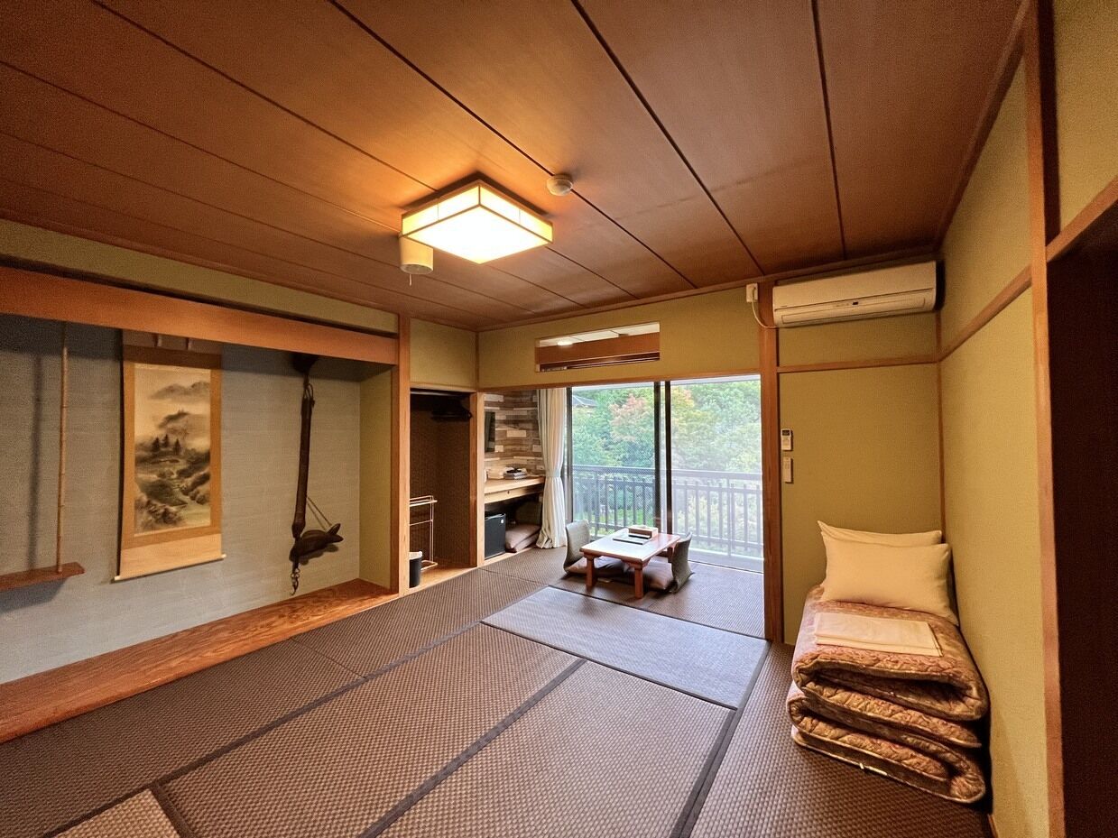 A traditional Japanese room with tatami flooring, a low table, a bed with a futon, a sliding door leading to a balcony, and a mounted air conditioner.