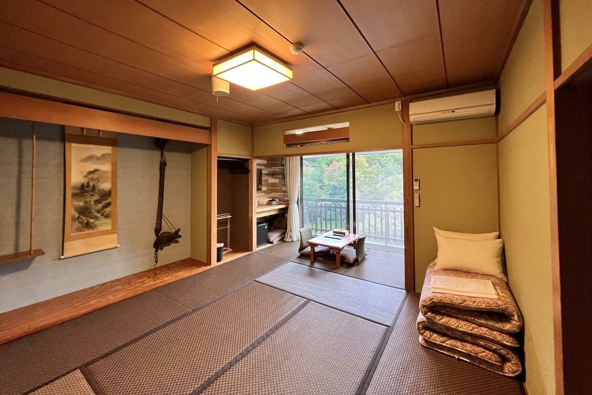 Ein traditioneller japanischer Raum mit Tatami-Bodenbelag, einem niedrigen Tisch, einem Bett mit Futon, einer Schiebetür zu einem Balkon und einer Wand-Klimaanlage.