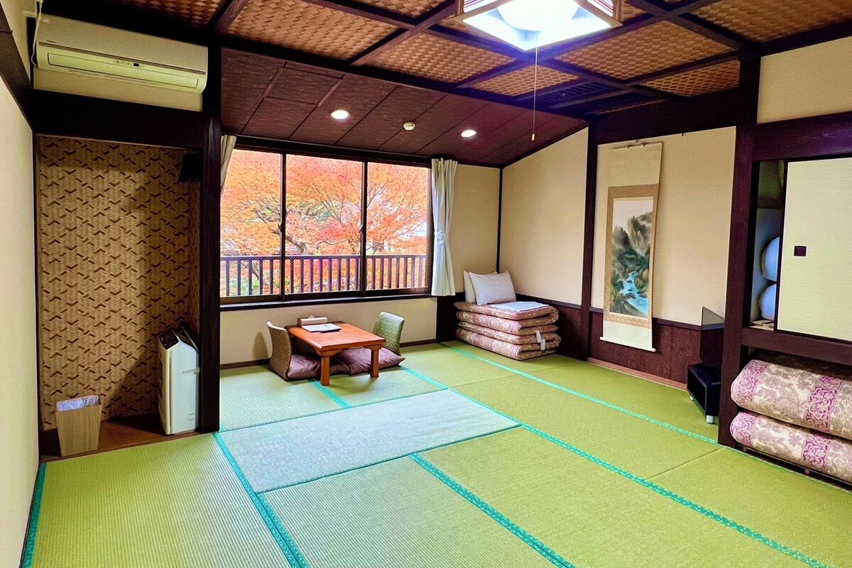 Ein traditioneller japanischer Raum mit Tatami-Boden, einem niedrigen Tisch, Kissen, einem Fenster mit Herbstlaub und einem Deckenventilator.