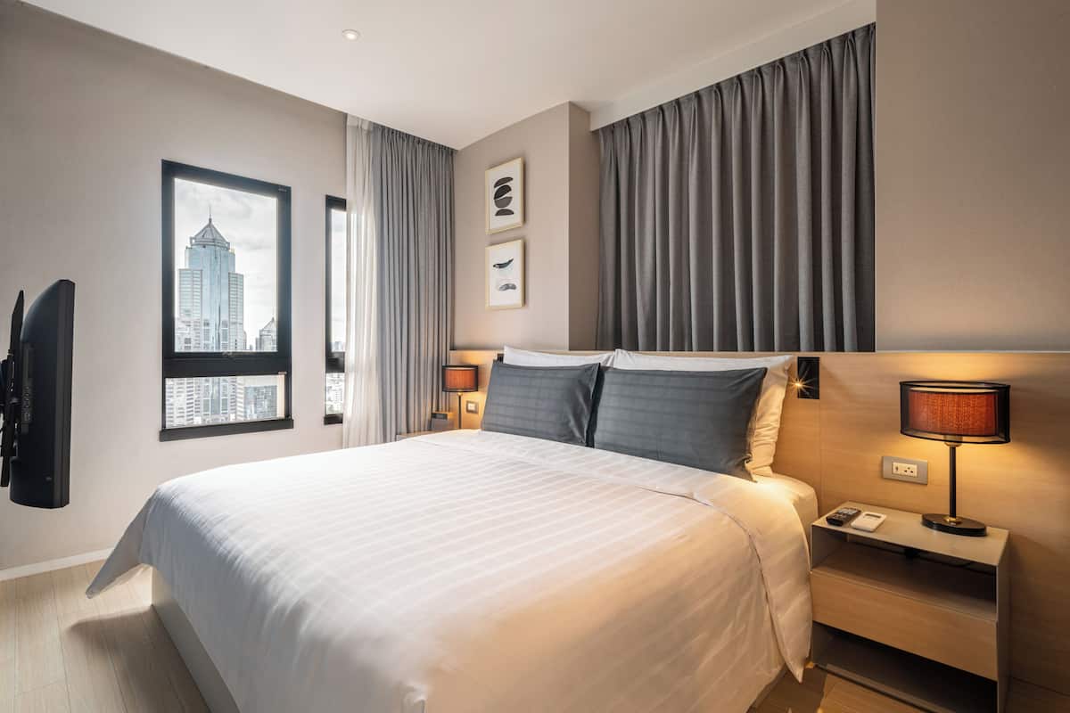 Una habitación de hotel moderna con una cama grande, vistas a la ciudad, un televisor de pantalla plana y una mesita de noche con lámpara.