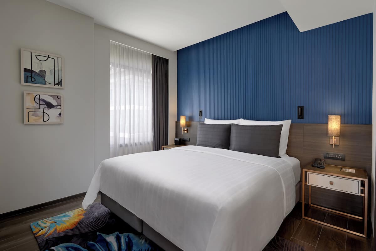Una habitación de hotel moderna con una cama grande, una mesita de noche con lámpara y arte abstracto en la pared.