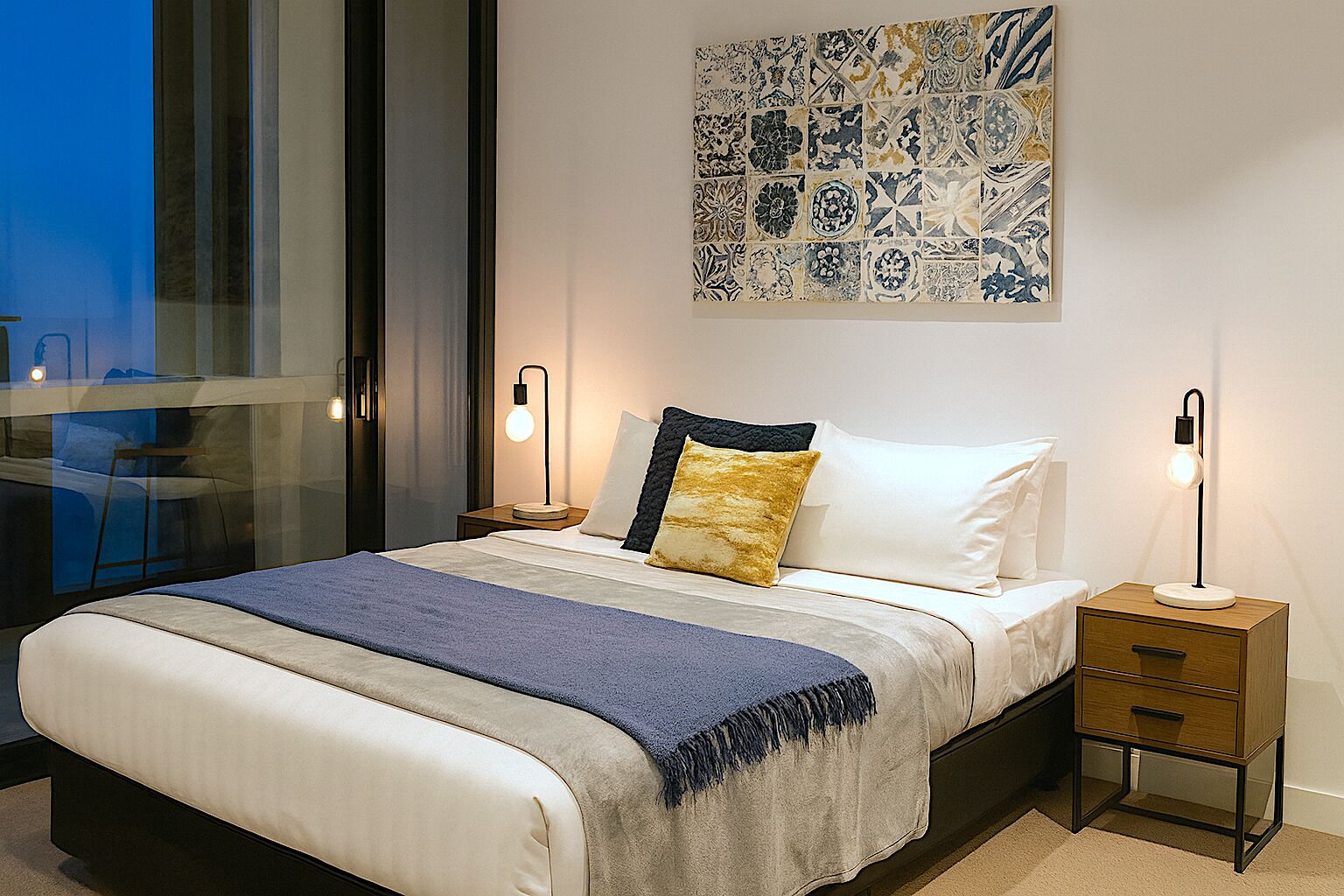 Un dormitorio moderno con una cama grande, mesitas de noche, una lámpara de noche, una obra de arte en la pared y vistas a través de una ventana.