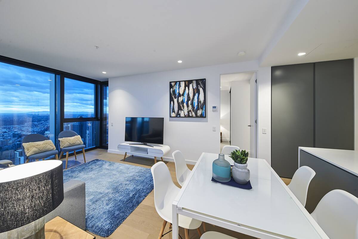 A modern living room with a large window, a flat-screen TV, a dining table, and a seating area with chairs.