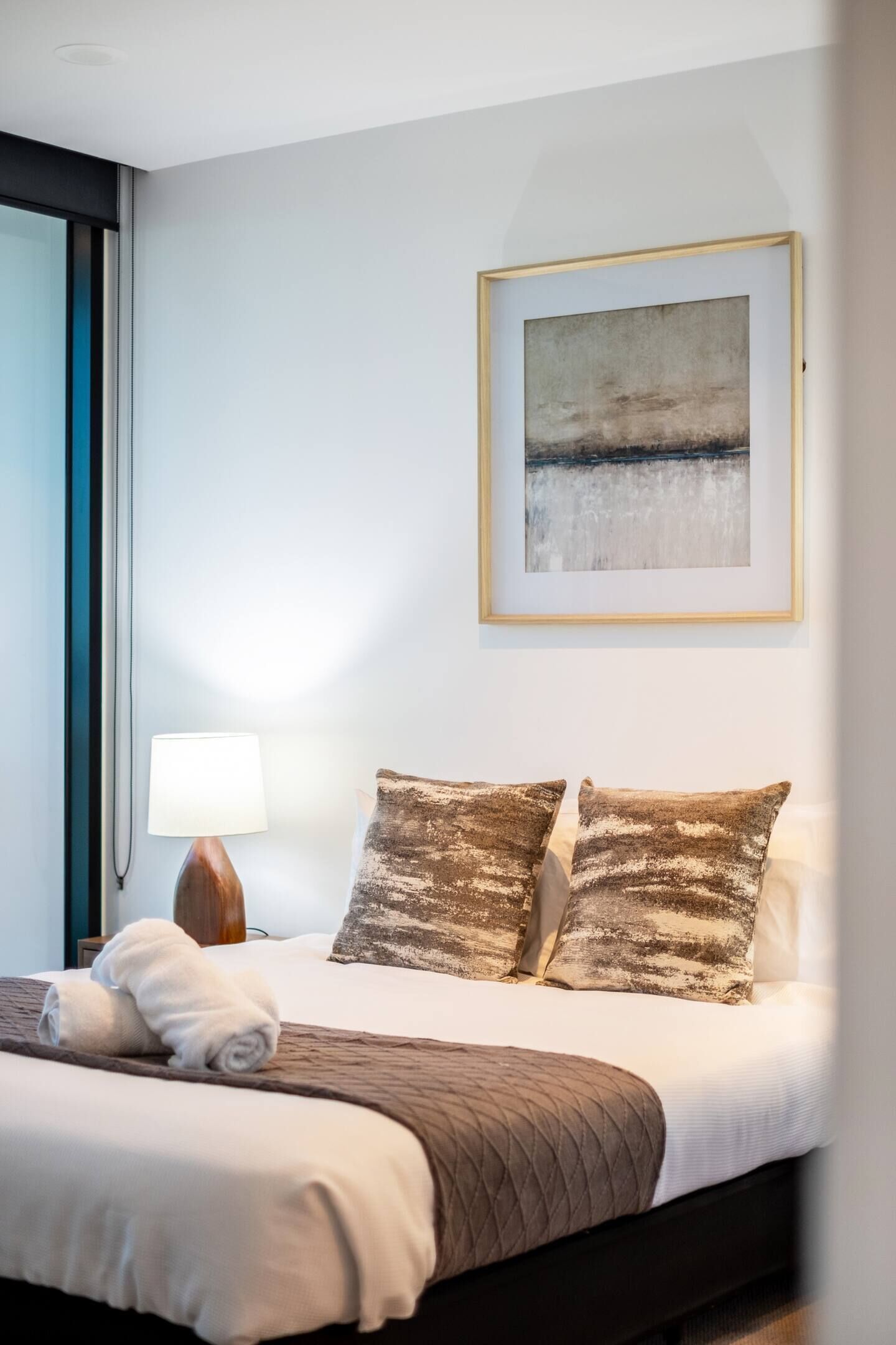 A neatly made bed with a quilted blanket, two pillows, a framed abstract painting, a bedside lamp, and a window with blinds.