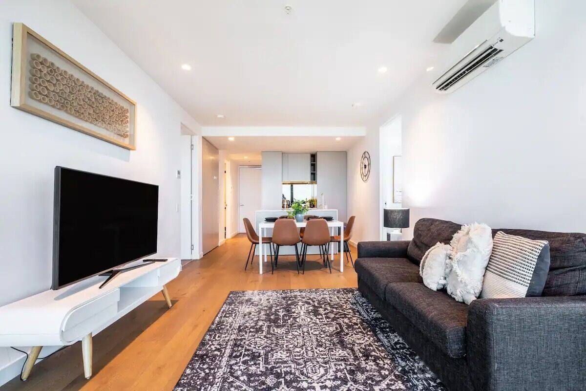 A modern living room with a flat-screen TV, a dark-colored sofa with decorative pillows, a patterned rug, and a dining area with a white table and chairs.