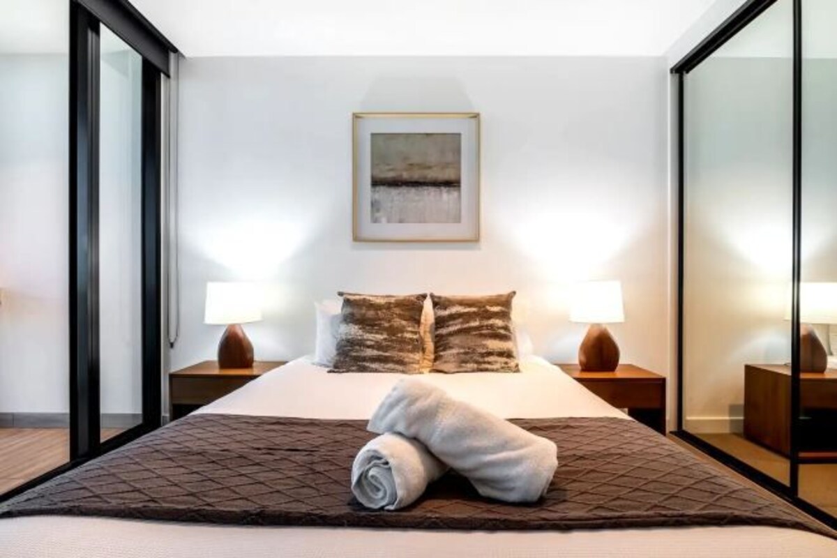 Une chambre d’hôtel moderne avec un grand lit, deux tables de chevet, un tableau au mur et une vue sur l’extérieur, visible à travers une porte vitrée.