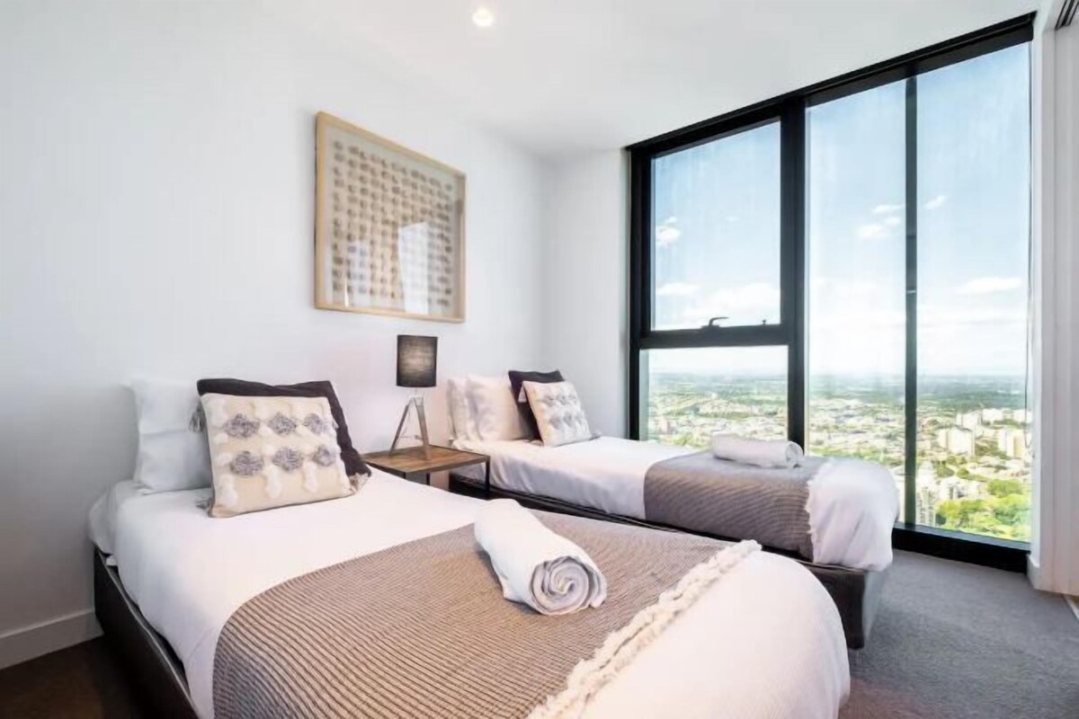 A hotel room with two beds, a large window with a city view, and a framed artwork on the wall.