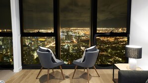 Una habitación moderna con un ventanal que ofrece vistas a la ciudad, dos sillas grises, una mesita y una lámpara.