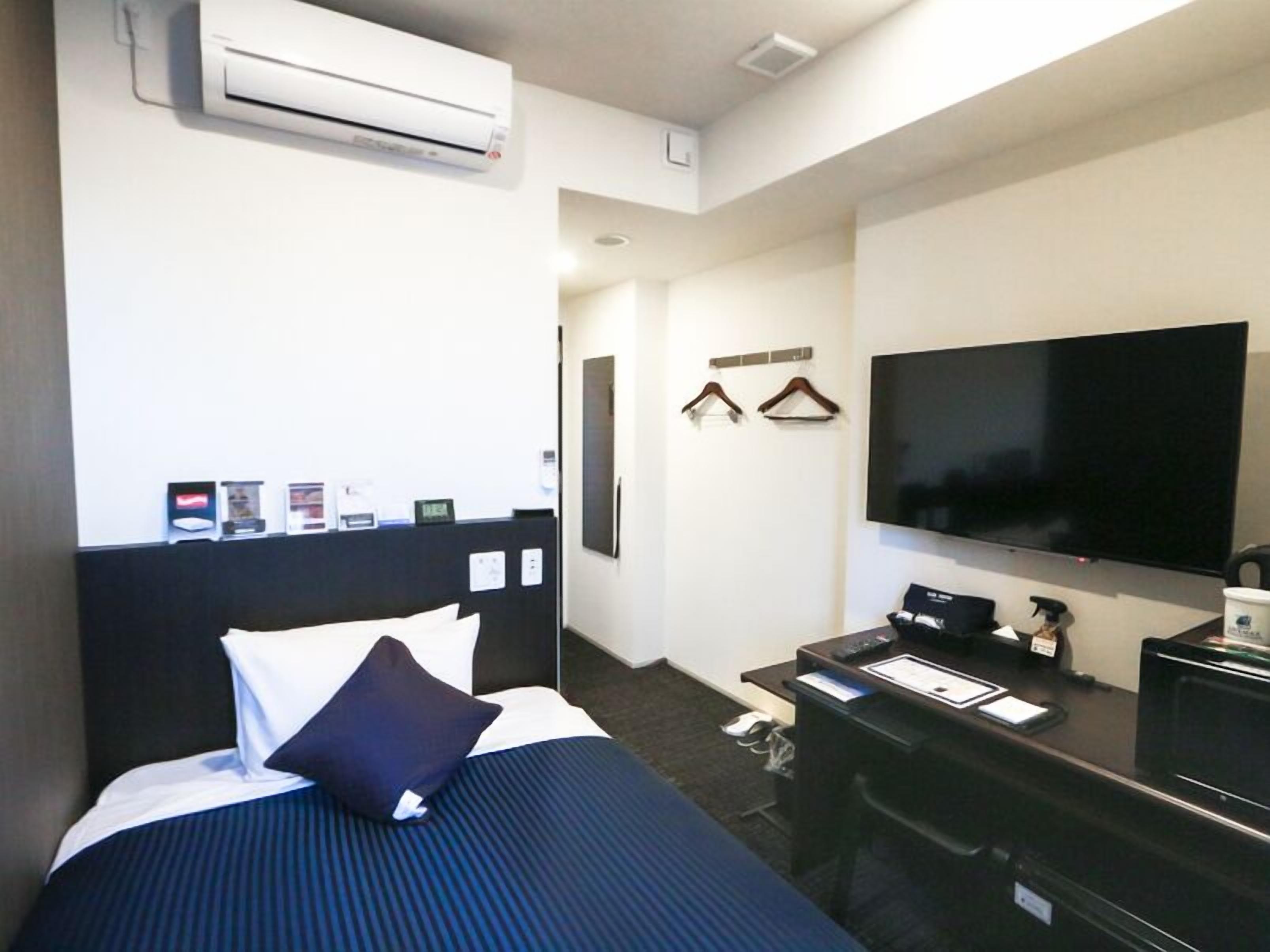 A hotel room with a bed, a desk, a flat-screen TV, and an air conditioning unit.