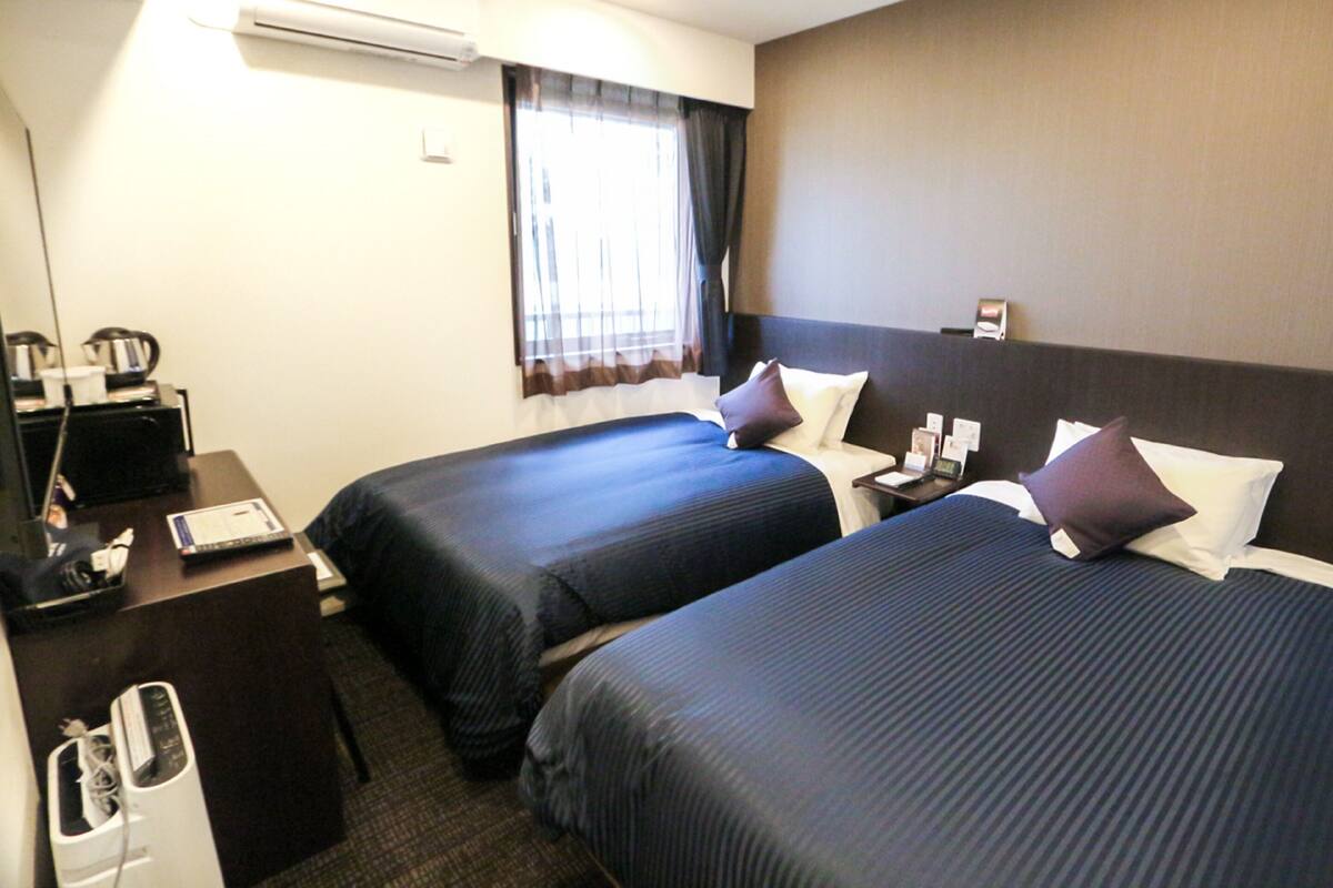 A hotel room with two beds, a desk, a chair, an air conditioner, and a window with curtains.