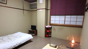 Una habitación pequeña y limpia con una cama, un escritorio con una computadora, una silla, un estante con recipientes rojos y una ventana con vistas.