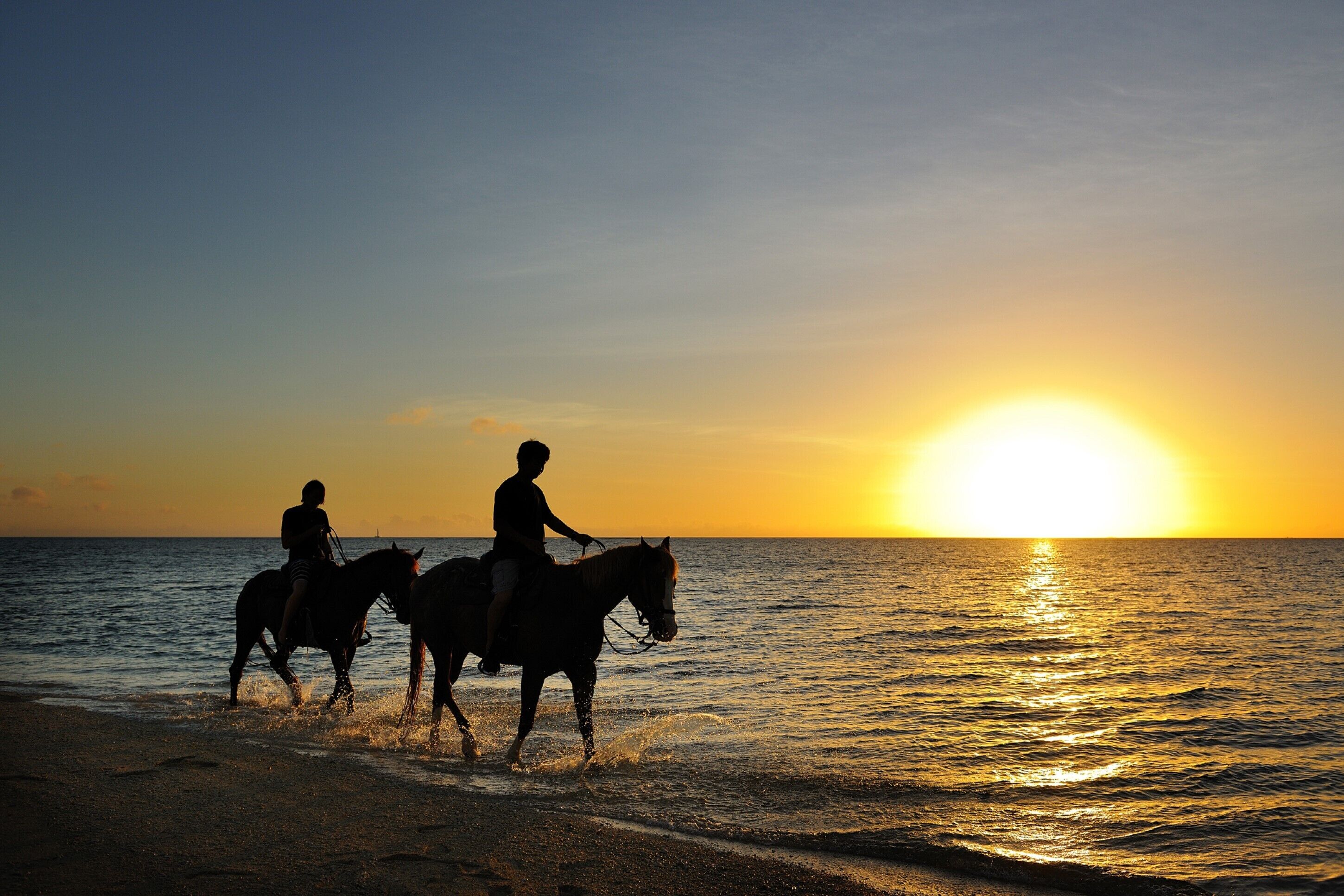 Twee mensen rijden op paarden langs een strand bij zonsondergang.