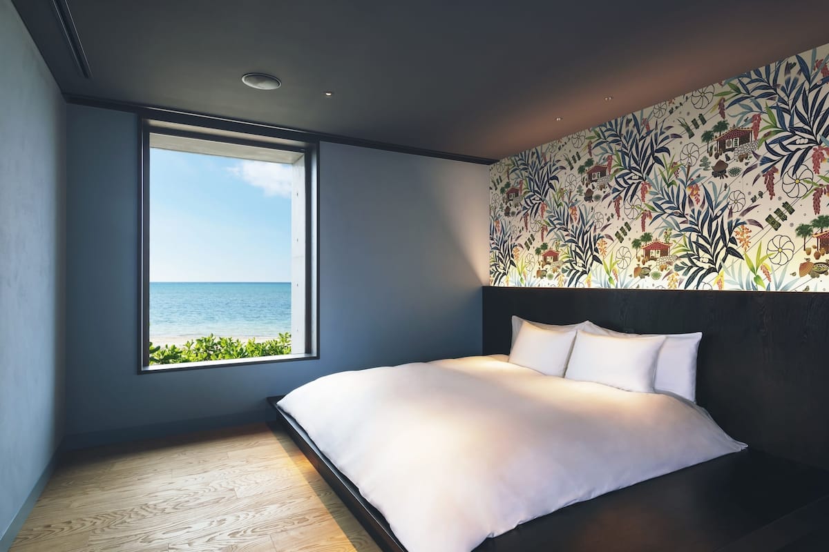 A bedroom with a large window overlooking the sea, a bed with white bedding, and a patterned wallpaper on the wall.