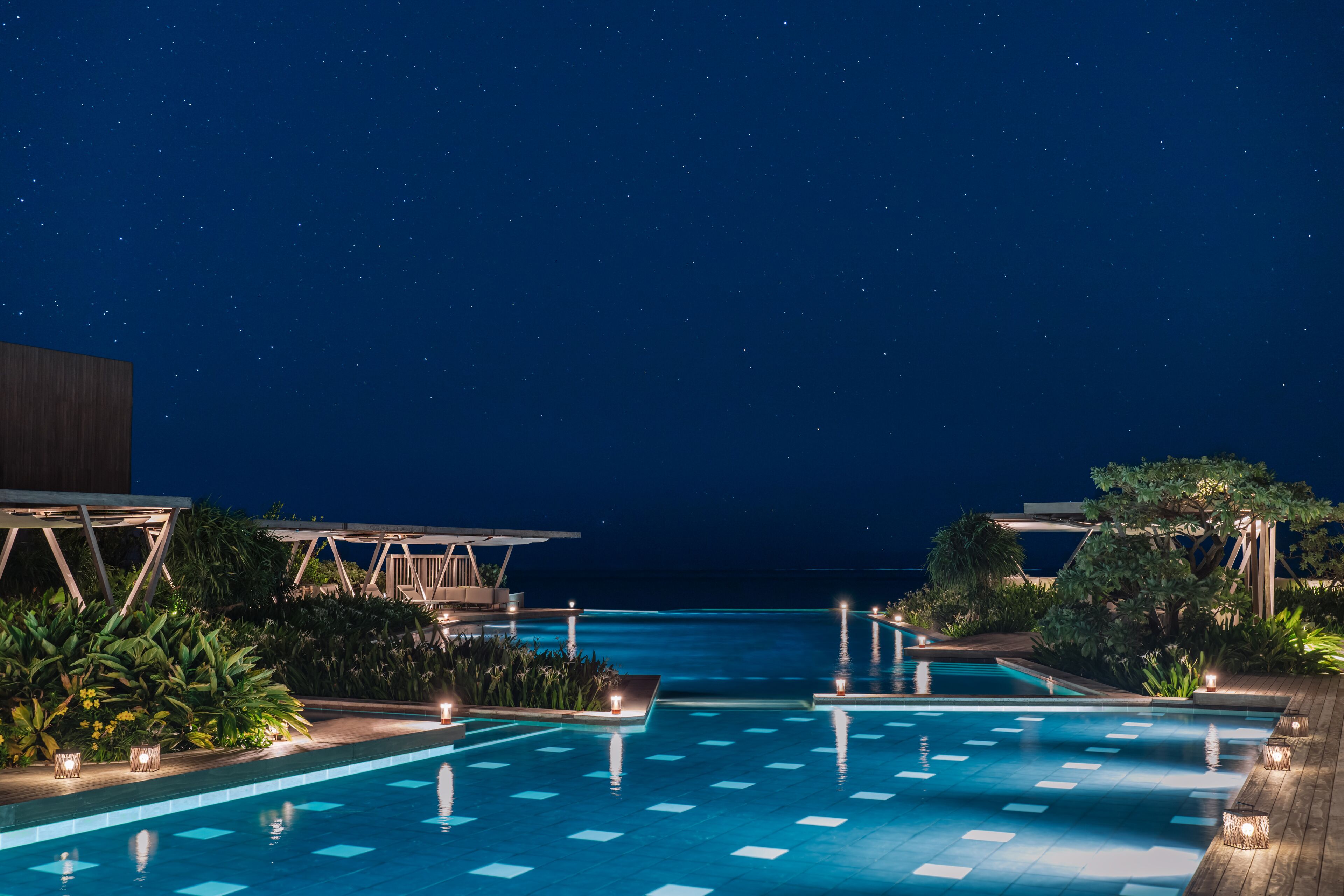 A nighttime swimming pool with a view of the ocean, surrounded by lush greenery and outdoor seating areas.