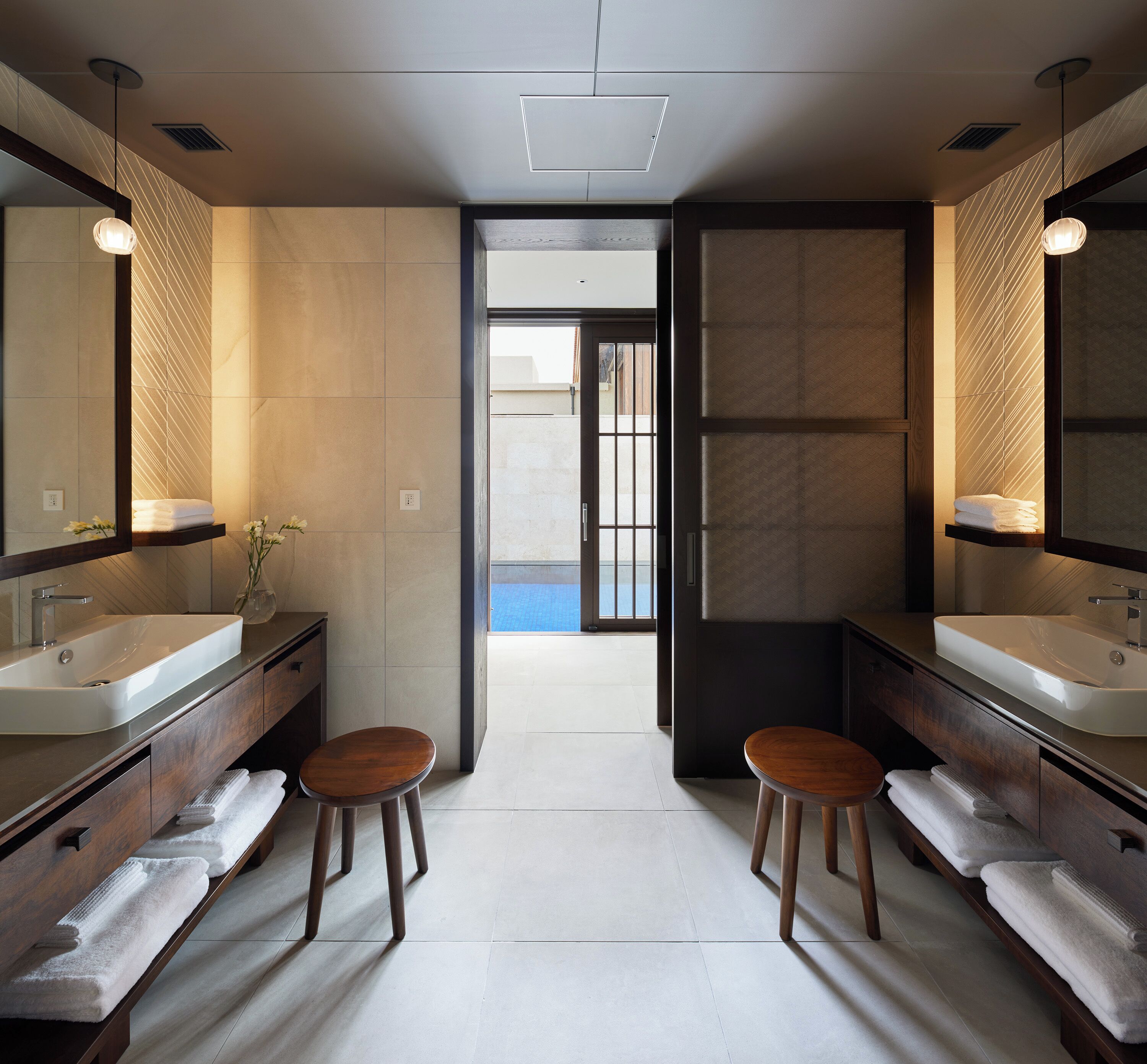 A modern bathroom with two sinks, wooden stools, and a large mirror.