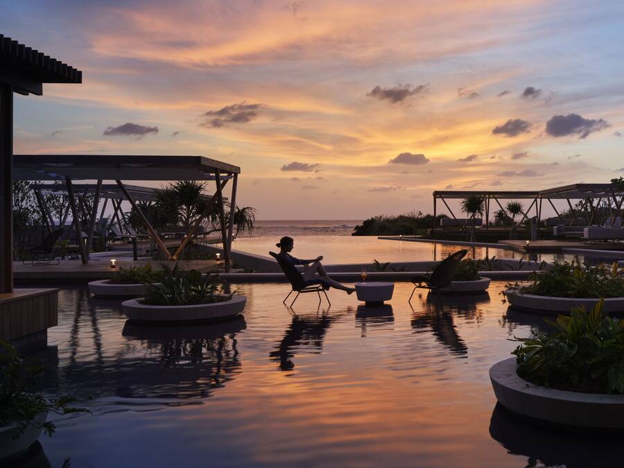 Un área de piscina tranquila con sillones de descanso y jardineras, con vista a un cuerpo de agua y un cielo al atardecer.
