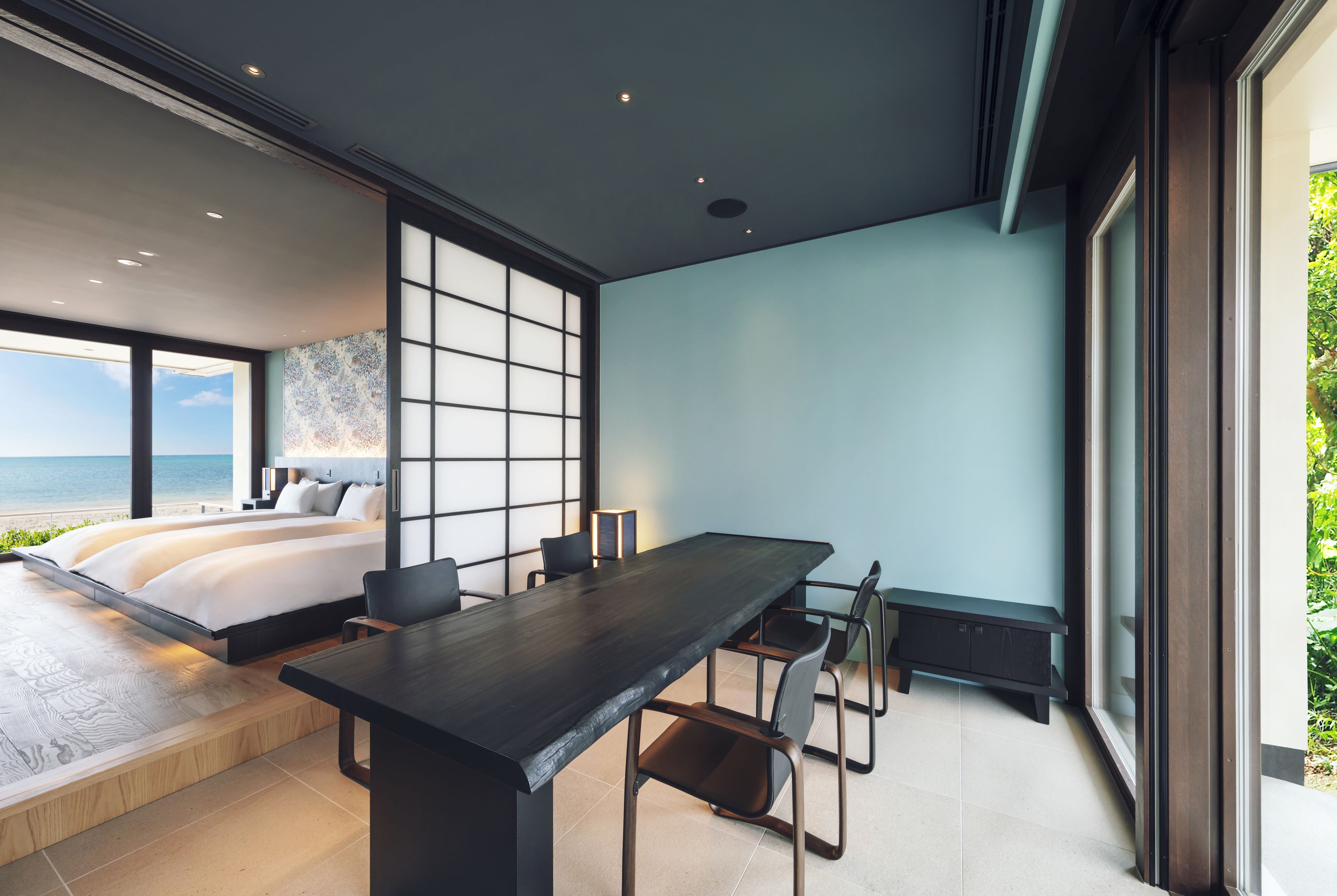 A modern bedroom with a large bed, a dining table, and a view of the sea.