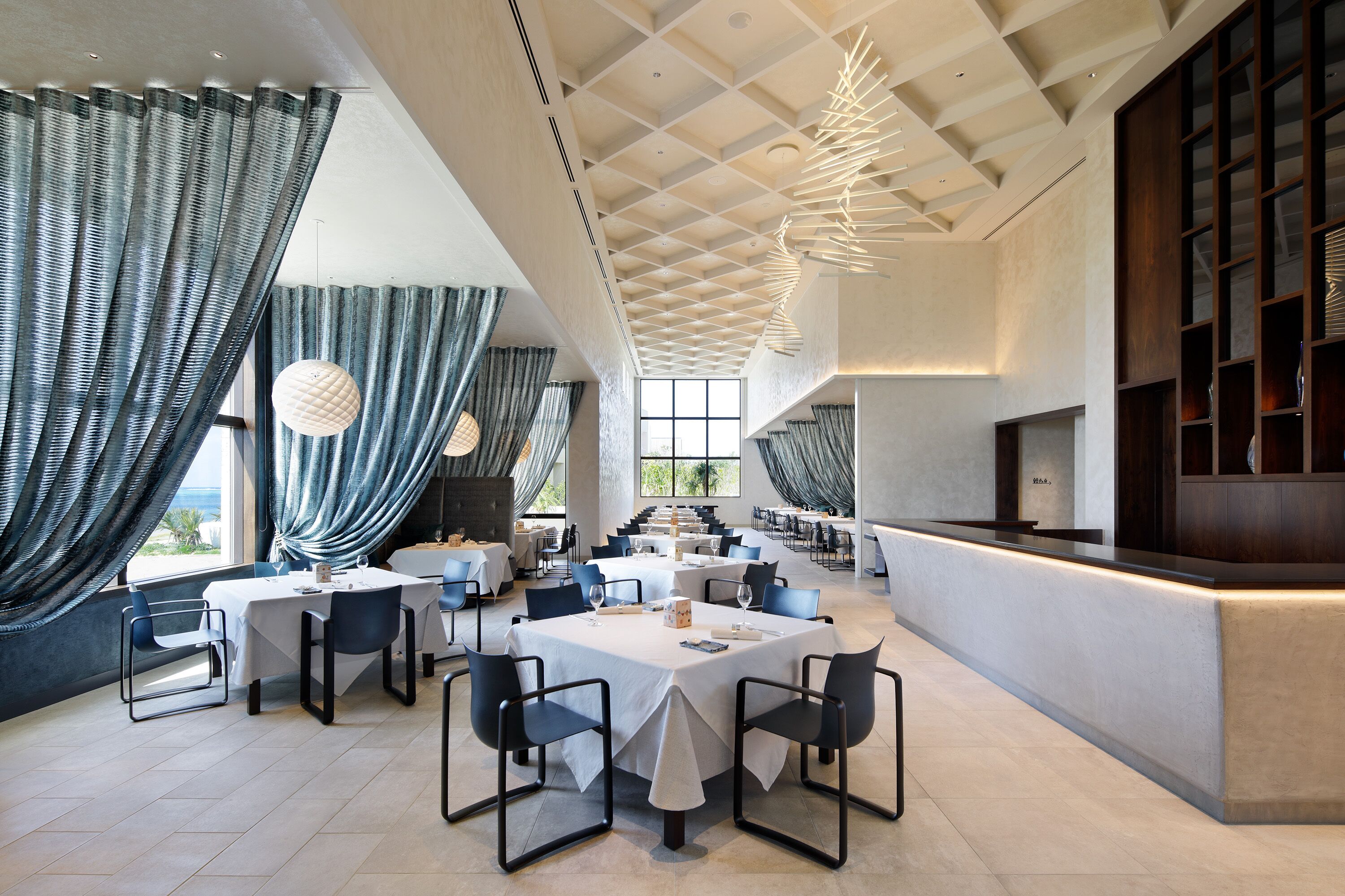 A spacious dining area with high ceilings and modern lighting fixtures. Tables are set with white tablecloths and dark chairs.