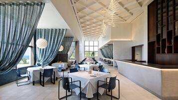 A spacious dining area with high ceilings and modern lighting fixtures. Tables are set with white tablecloths and dark chairs.