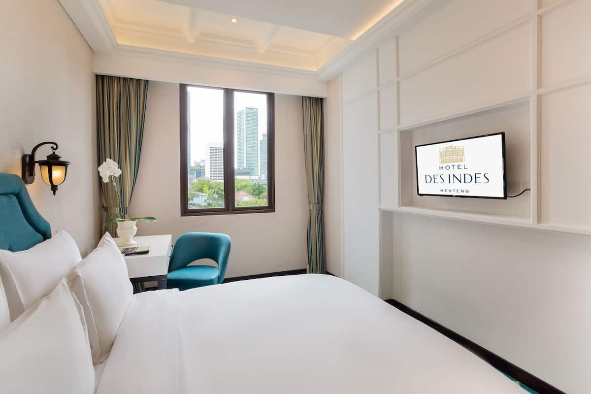 A hotel room with a large bed, a desk, a chair, a TV mounted on the wall, and a window with a city view.