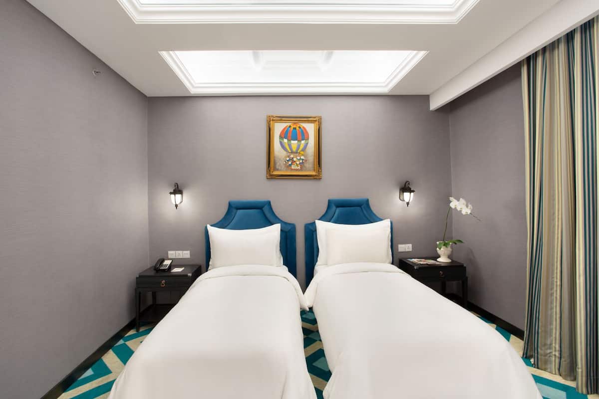 A hotel room with two beds, a skylight, a painting, and a nightstand with a phone and a vase of flowers.
