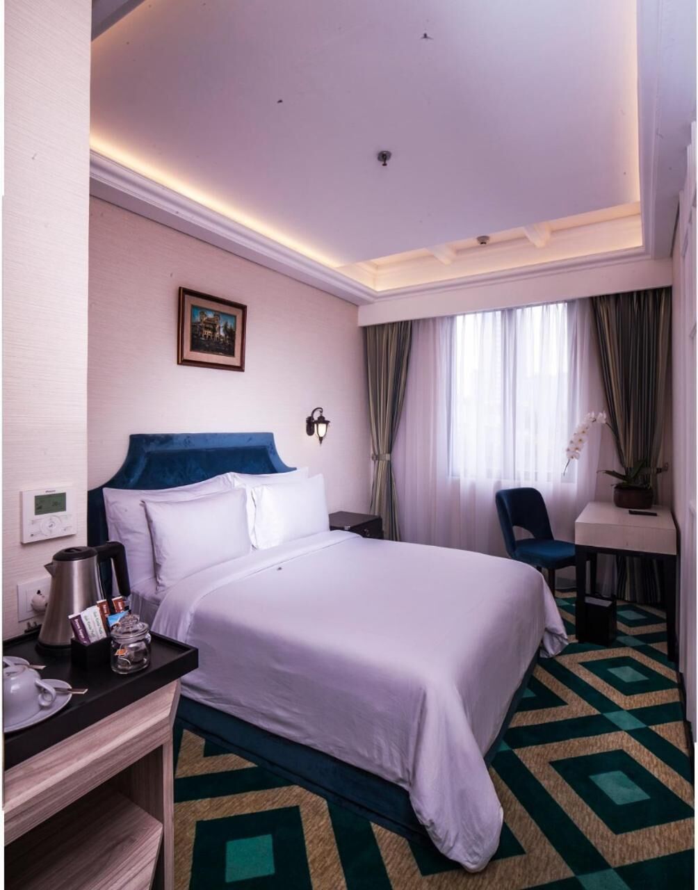 Una habitación de hotel con una cama grande, un escritorio con una silla, una mesita con una planta en maceta y un cuadro en la pared.