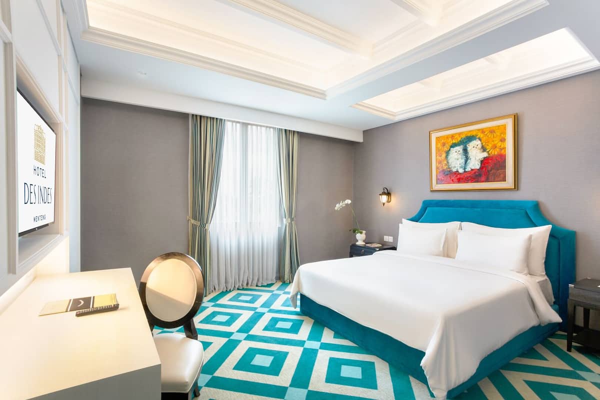 A hotel room with a large bed, a desk with a chair, a desk lamp, a mirror, and a painting on the wall.