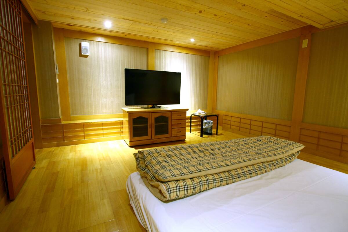 A hotel room with a wooden floor, a bed with a plaid blanket, a television on a wooden cabinet, and a window with blinds.