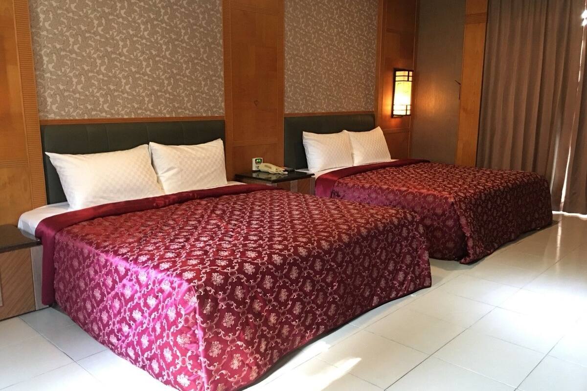 A hotel room with two beds, a nightstand, and a wall-mounted lamp.
