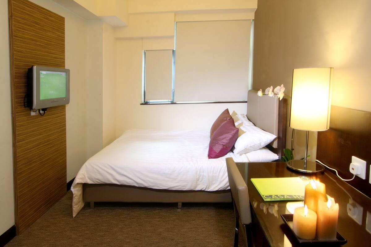 A hotel room with a bed, a desk with a lamp, a television, and a window.