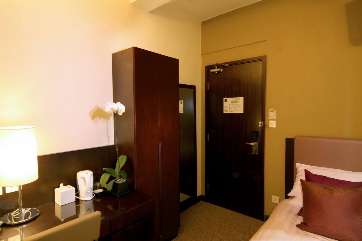 A hotel room with a bed, a desk with a lamp, a plant, and a door with a sign.