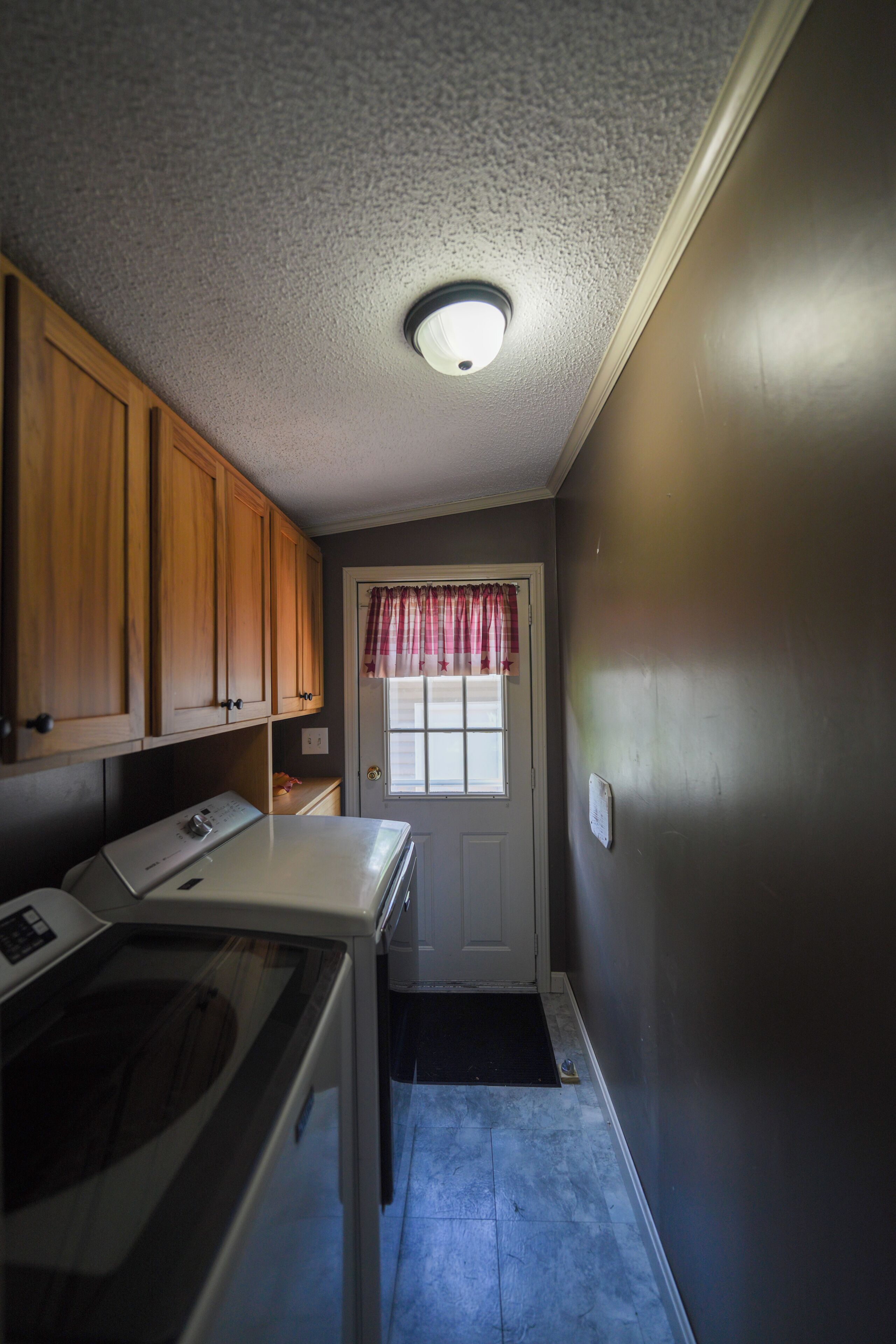 A laundry room with a washing machine, dryer, wooden cabinets, a window with a curtain, and a door.