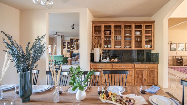 A dining area with a wooden table set for a meal, surrounded by chairs. There is a kitchen with wooden cabinets in the background.
