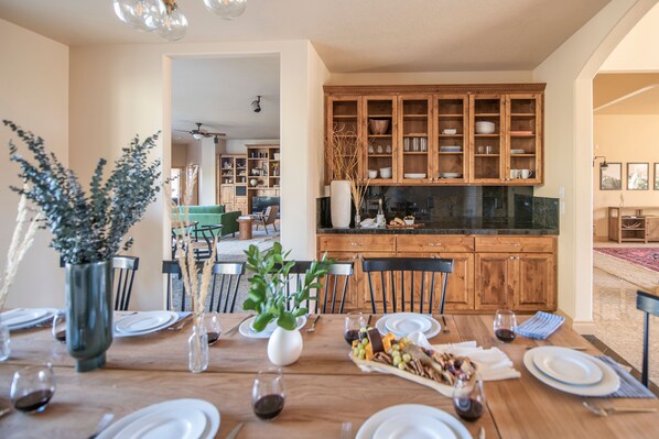 A dining area with a wooden table set for a meal, surrounded by chairs. There is a kitchen with wooden cabinets in the background.