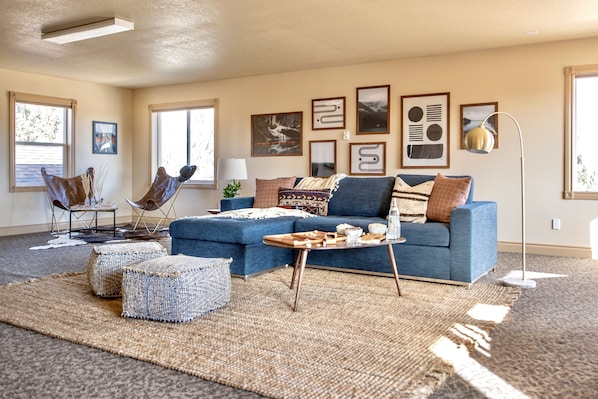 Ein Wohnzimmer mit einem blauen Sofa, einem Couchtisch und einem Teppich. An den Wänden hängen Bilder in Rahmen und eine Lampe steht bereit.