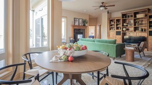 A dining area with a round table, chairs, and a bowl of fruit. There is a green sofa and a television in the background.