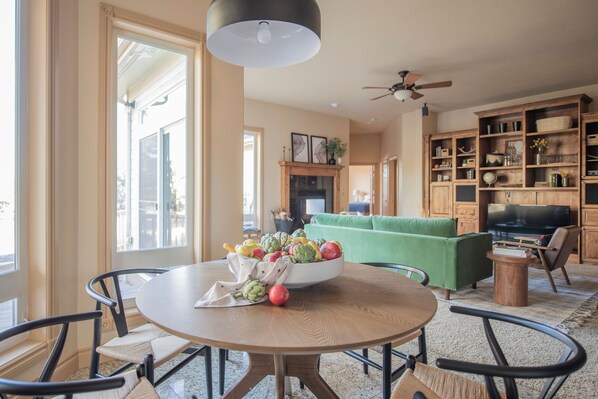 Un comedor con una mesa redonda, sillas y un frutero. Se observa un sofá verde y un televisor al fondo.