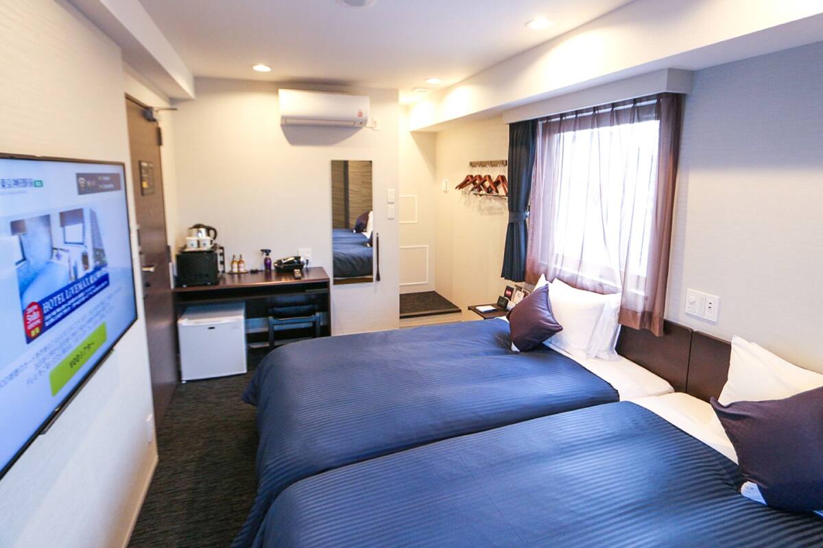 Habitación de hotel con dos camas, un escritorio, una silla, una kitchenette pequeña y una ventana con cortinas.