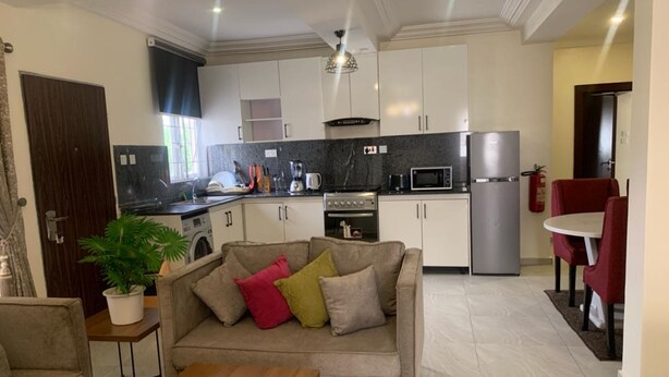 A modern kitchen with white cabinets, a microwave, and a refrigerator. There is a dining area with a red table and chairs. A beige sofa with colorful cushions is in the living area. - Tennys Place Apartments GARKI (Abuja)